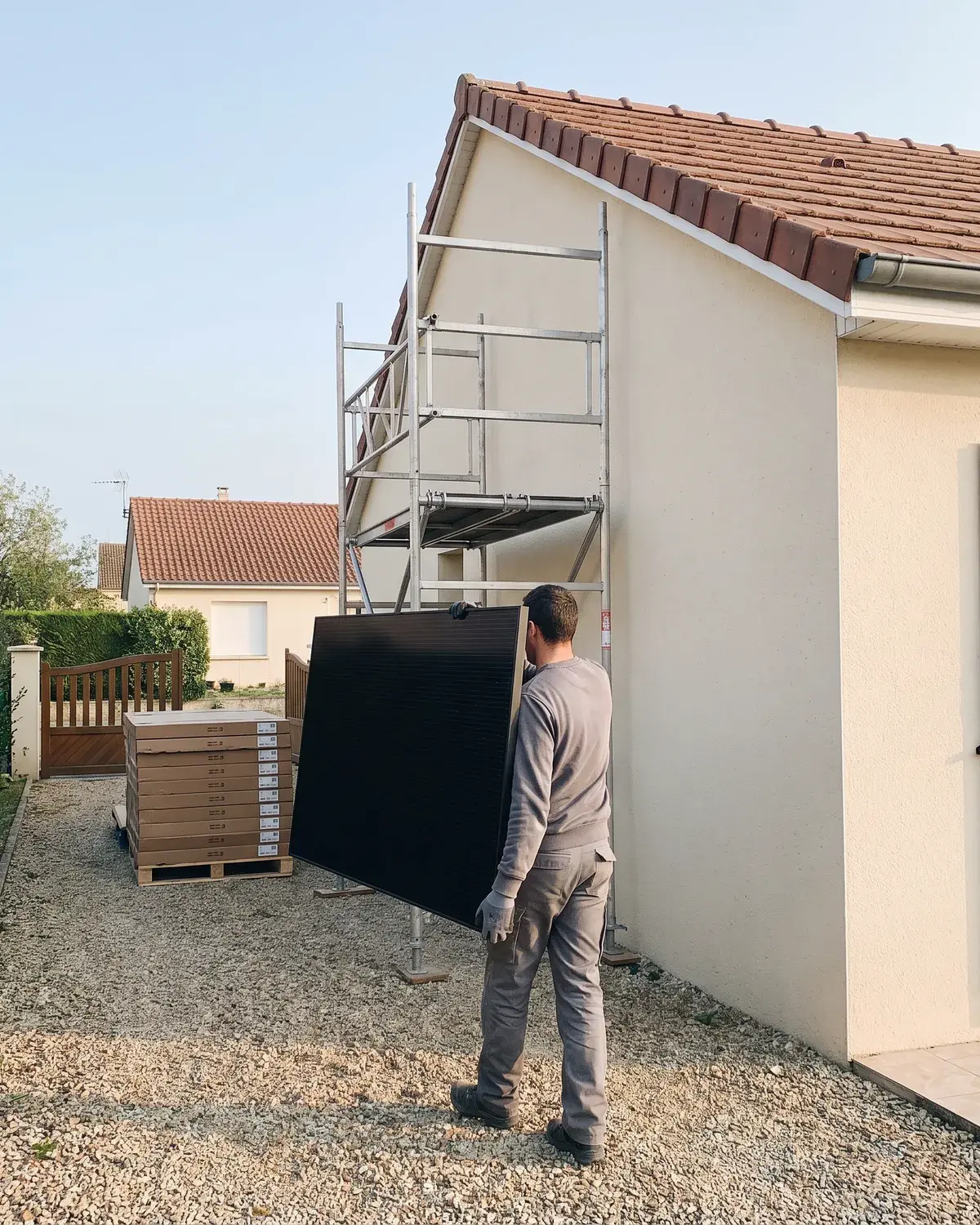 Un installateur porte un panneau LONGi vers l'échafaudage d'un pavillon, palette de modules au portail